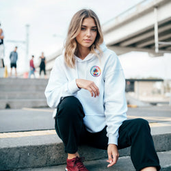Woman crouching in light blue hoodie with circular tropical palm tree graphic, black pants, and red sneakers in urban setting.