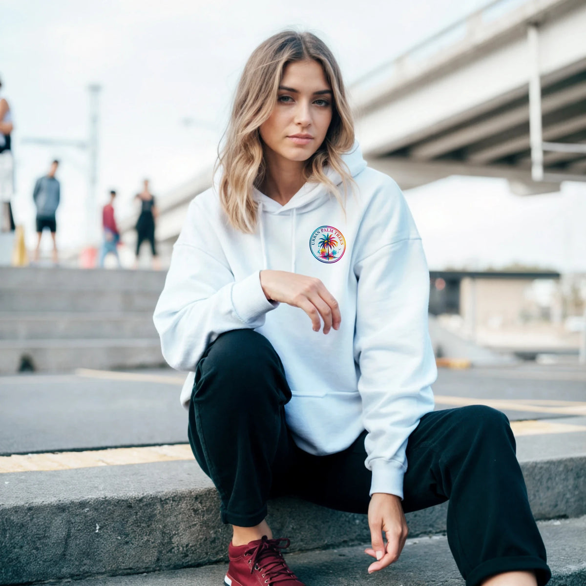 Woman crouching in light blue hoodie with circular tropical palm tree graphic, black pants, and red sneakers in urban setting.