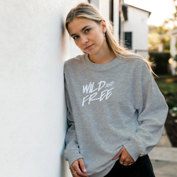 Woman wearing Cute Wild And Free Sweatshirt in gray with white script slogan, outdoors.