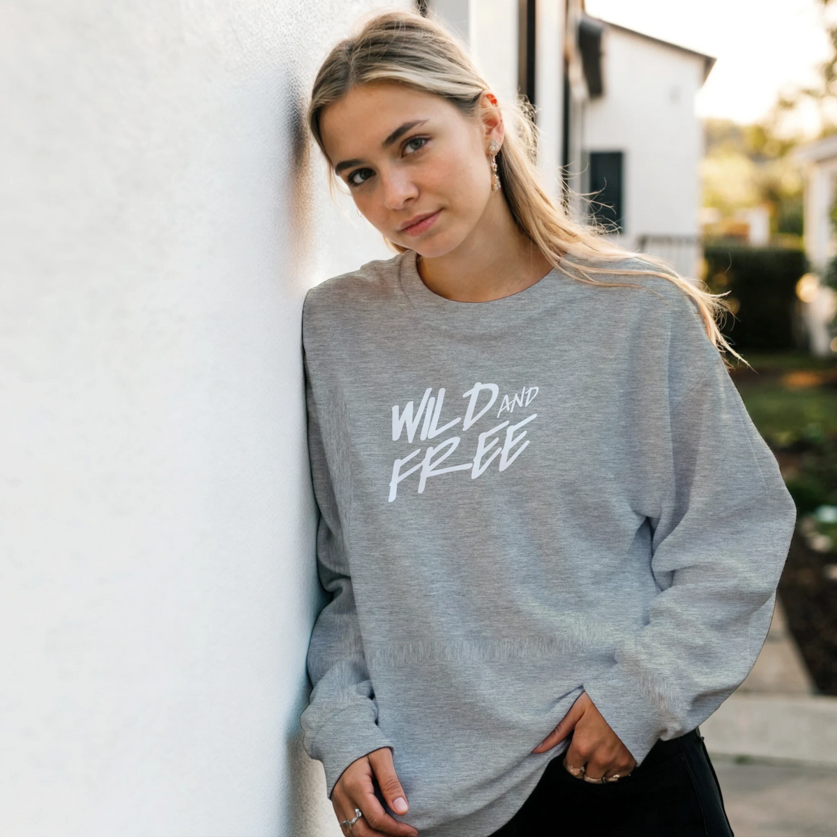 Woman wearing Cute Wild And Free Sweatshirt in gray with white script slogan, outdoors.