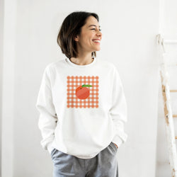Smiling woman in a white sweatshirt with orange gingham check and a peach graphic on the chest.