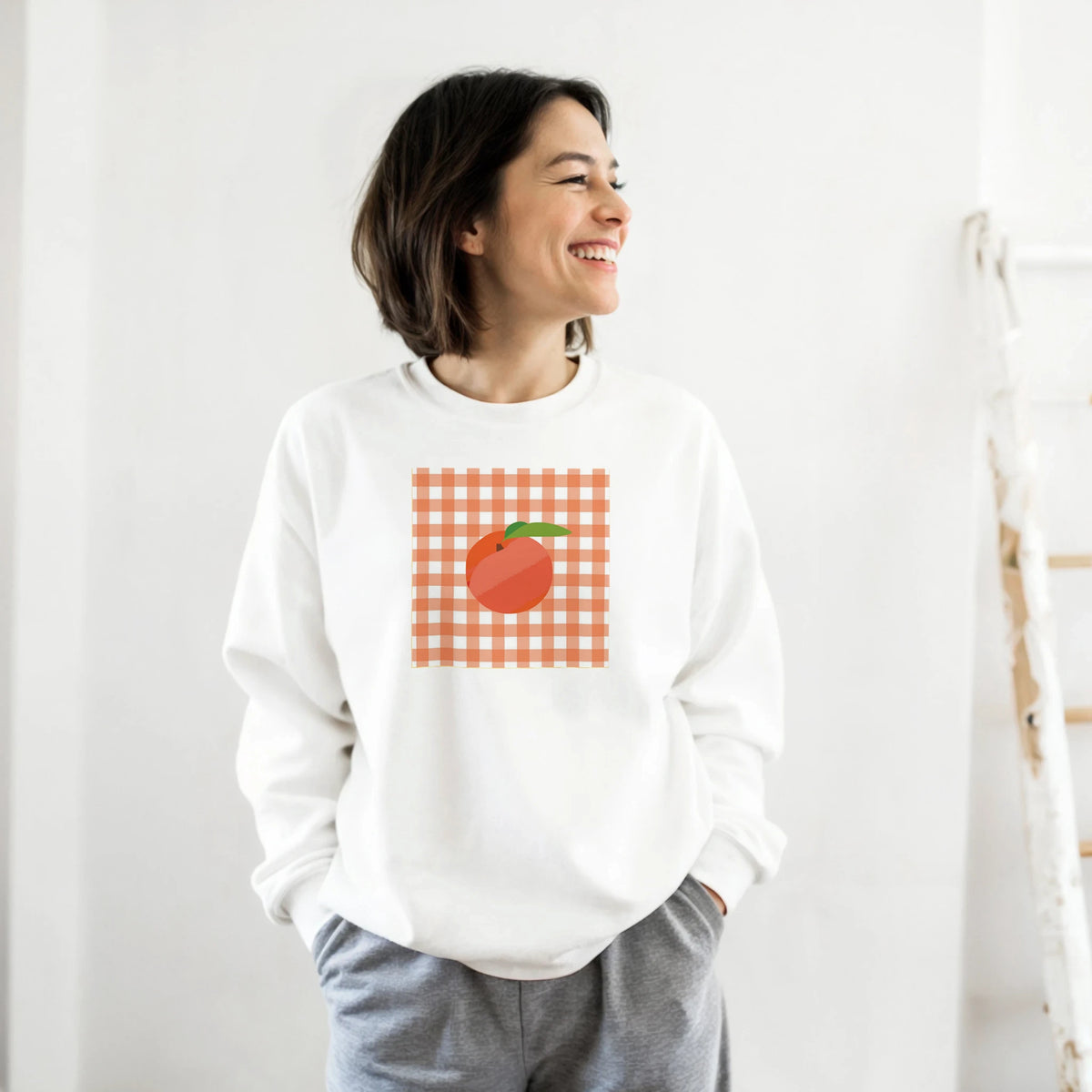 Smiling woman in a white sweatshirt with orange gingham check and a peach graphic on the chest.