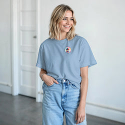 Woman wearing a light blue retro graphic t-shirt with a small palm tree chest logo, paired with high-waisted denim jeans.