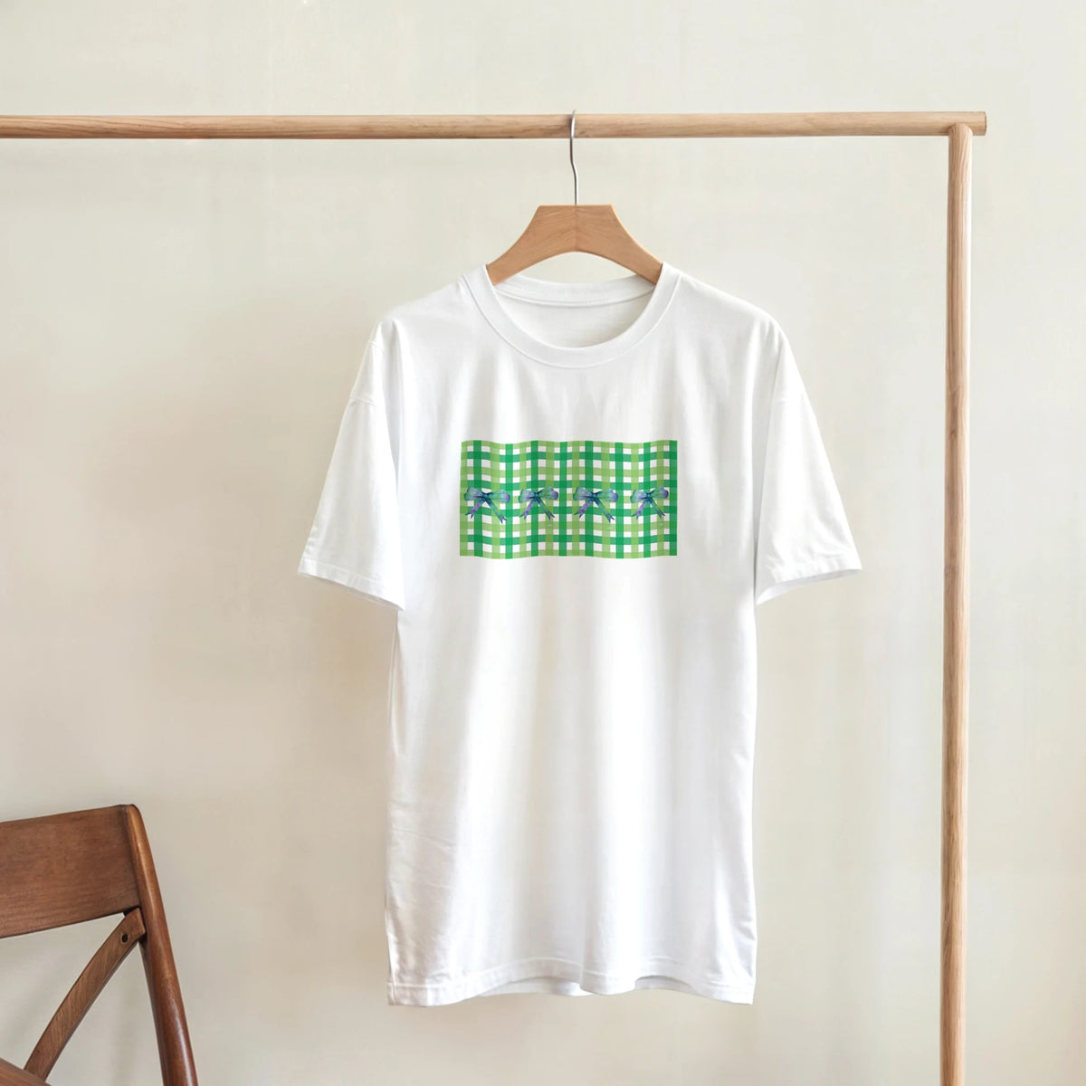 White women's T-shirt with a green gingham bow print on front, hanging on a wooden hanger.