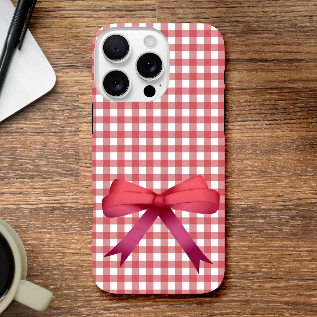 Red gingham iPhone case with a large pink bow on a wooden desk. Cute coquette tough phone cover for iOS 13-16.