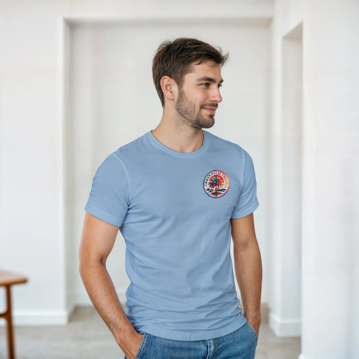 Man wearing light blue retro t-shirt with circular palm tree graphic on chest, styled casually with blue jeans in a bright room.