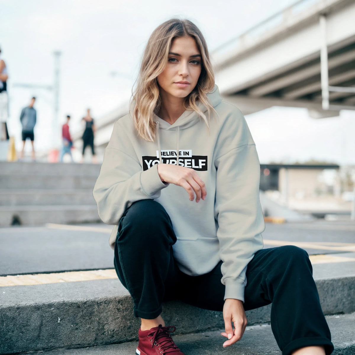 Woman in a light gray Believe In Yourself hoodie with bold front slogan, seated on urban steps.