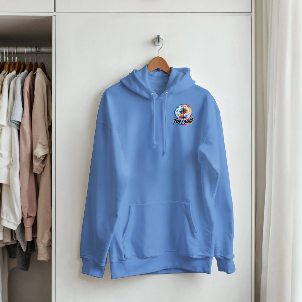 Light blue streetwear hoodie with palm tree graphic, front pouch pocket, and drawstrings hanging in a closet.