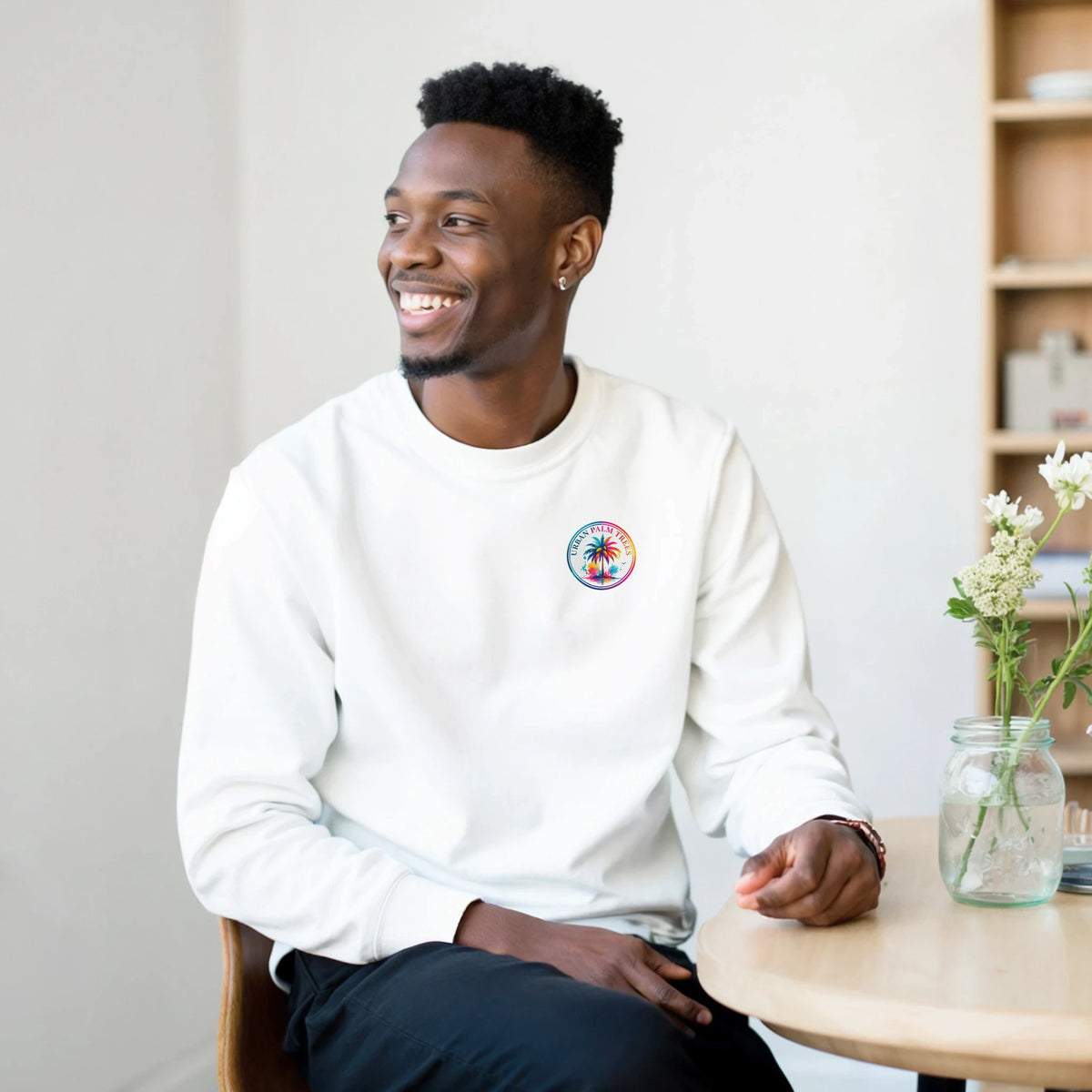 Man wearing a white crew neck sweatshirt featuring a small, colorful circular Urban Palm Trees logo on the left chest.