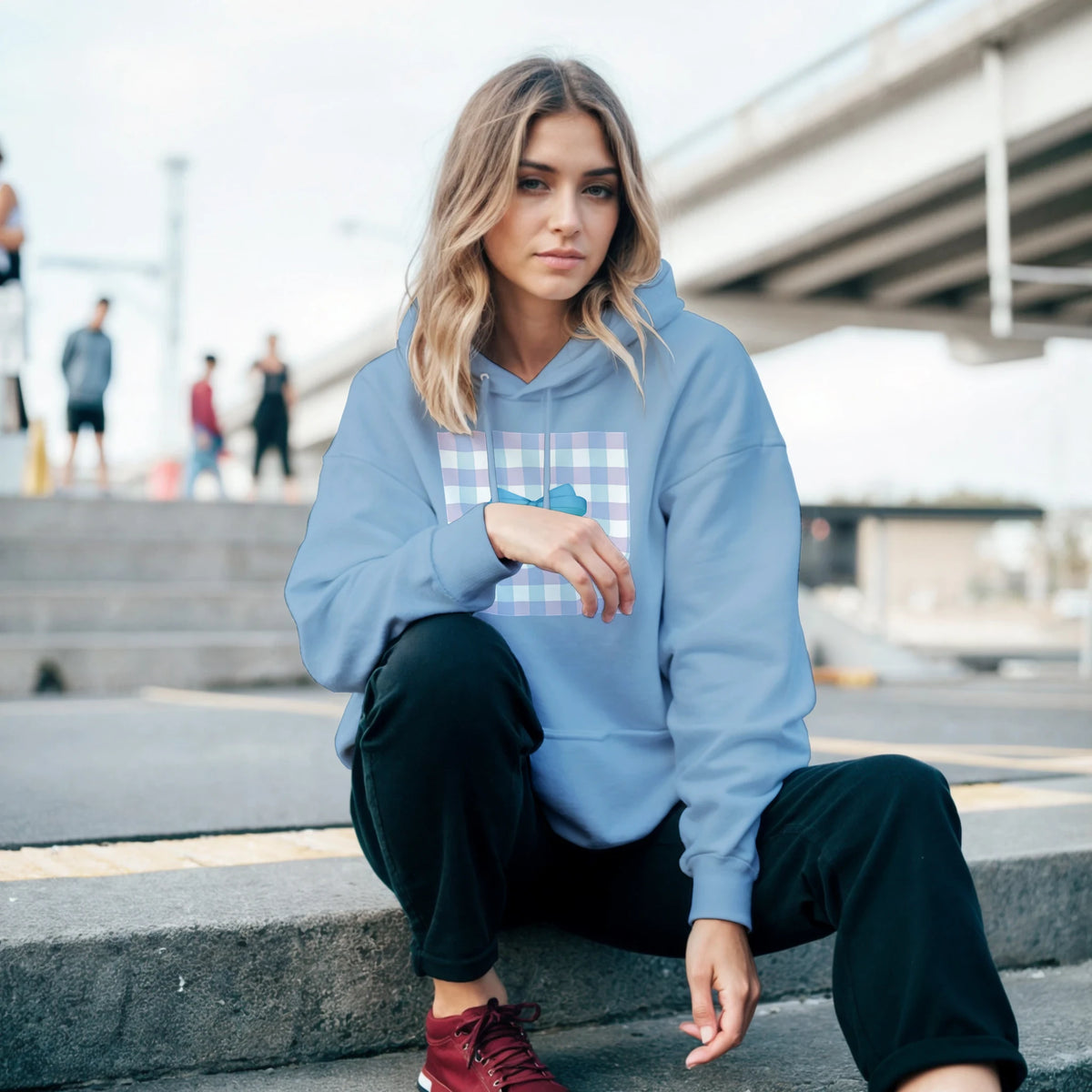 Woman sitting on steps wearing a light blue coquette graphic pullover hoodie featuring a plaid bow design and relaxed fit.