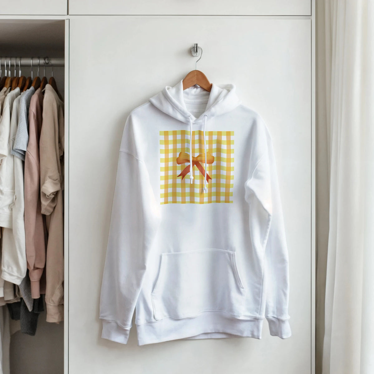 White hoodie with yellow gingham bow graphic hanging on a wooden hanger in a closet.