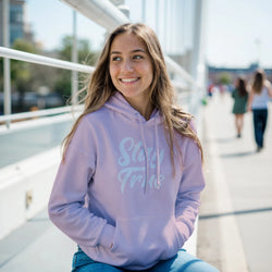 Smiling woman in a lavender Stay True hoodie with front pouch pocket, outdoors in an urban setting.