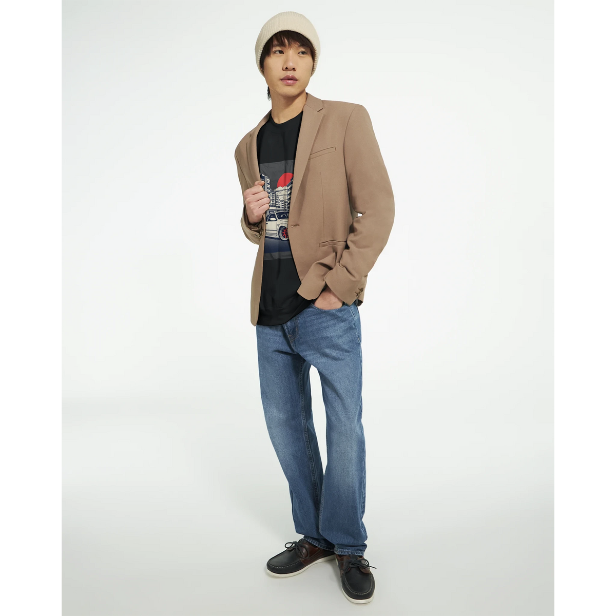 Male model wearing tan blazer, black graphic T-shirt, blue jeans, and beige beanie.