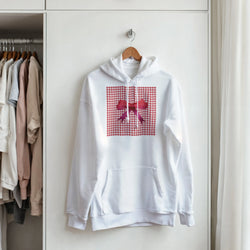 White women's hoodie with red gingham panel and pink bow, hanging on a hanger in a closet.