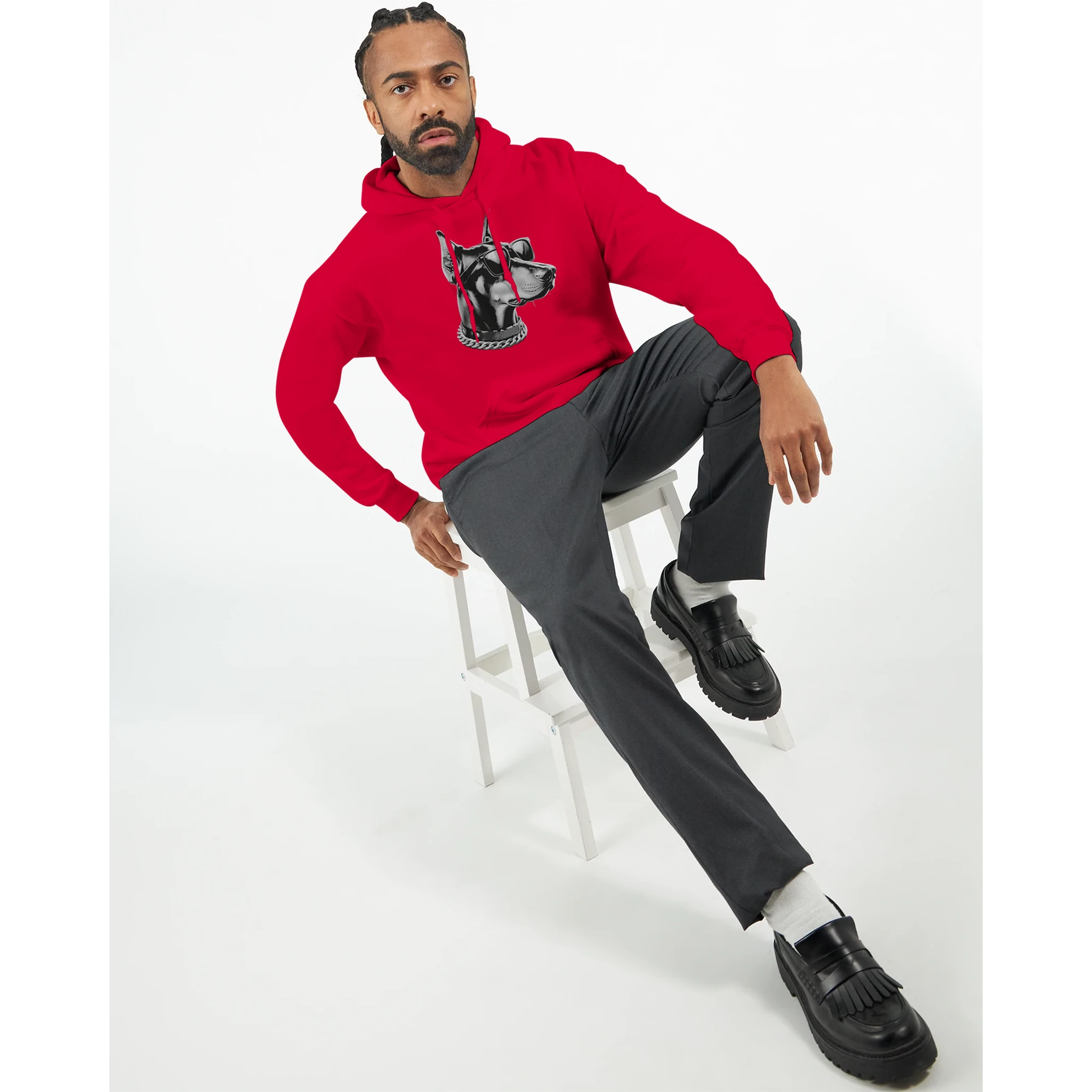 Man wearing red Gangster Dog Jumper graphic hoodie, seated on white stool, black loafers.