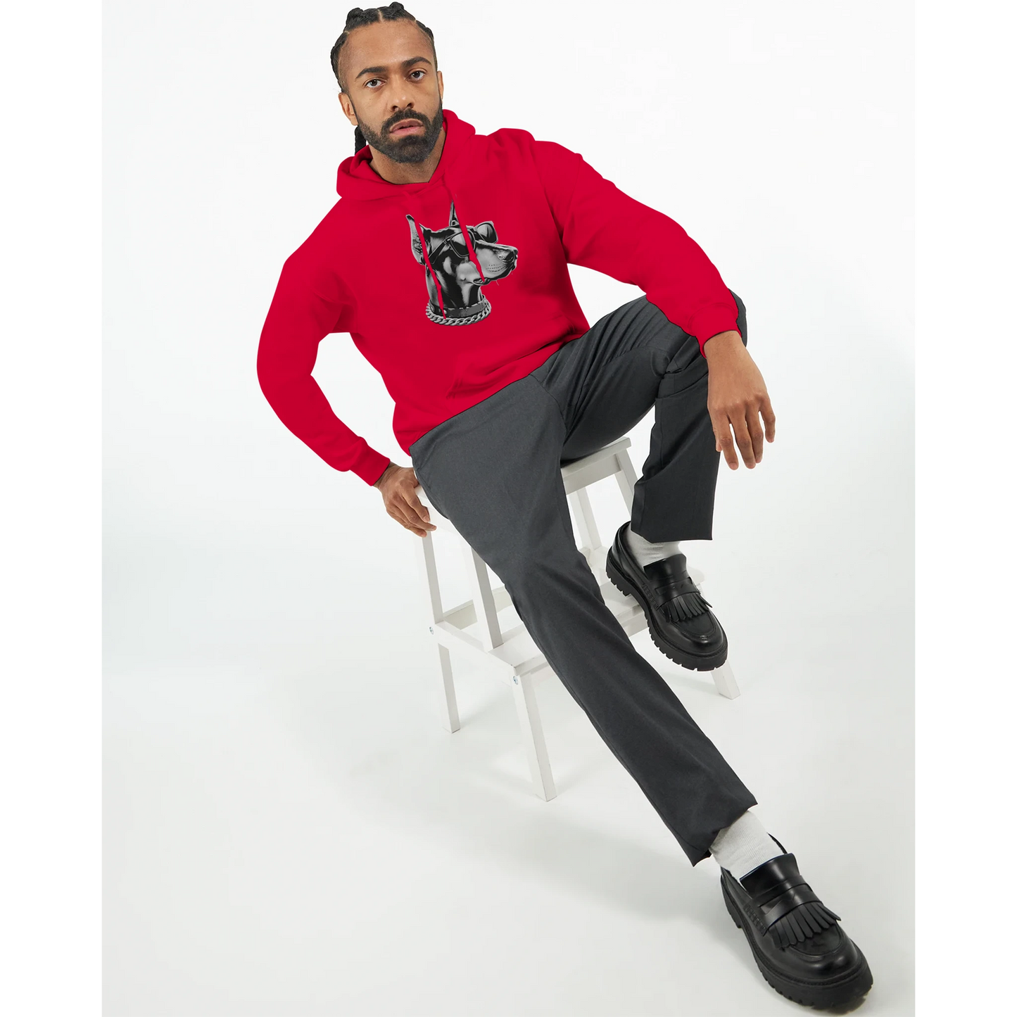 Man wearing red Gangster Dog Jumper graphic hoodie, seated on white stool, black loafers.