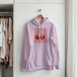 Lavender retro hoodie featuring a red cherry graphic on a gingham patch, hanging in a closet.