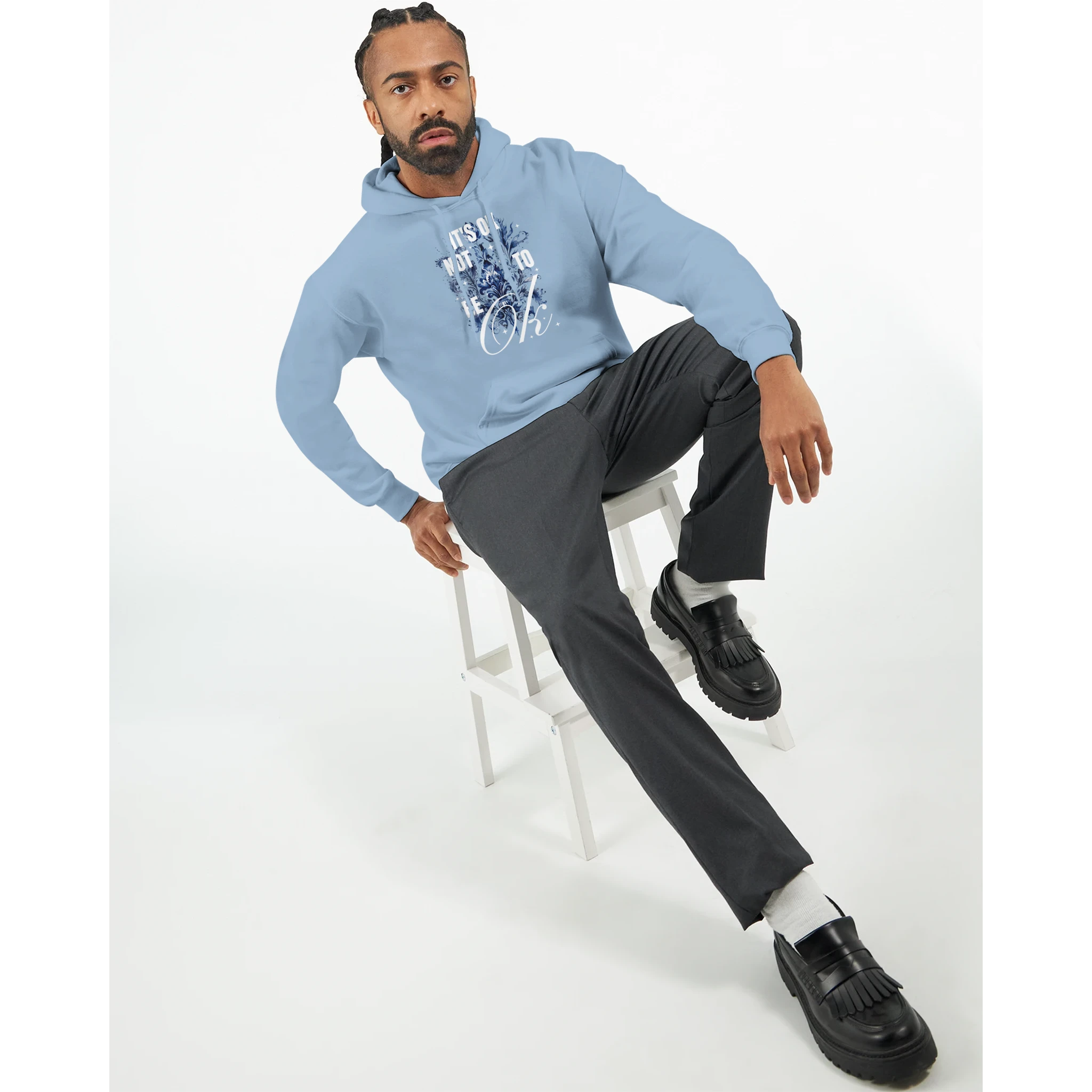 Man wearing light blue cotton-blend hoodie with 'It's OK Not To Be OK' graphic, seated on a white stool.