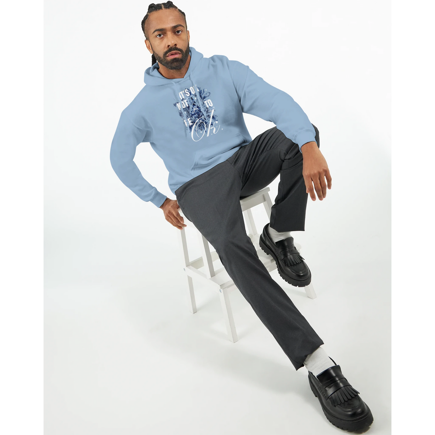 Man wearing light blue cotton-blend hoodie with 'It's OK Not To Be OK' graphic, seated on a white stool.