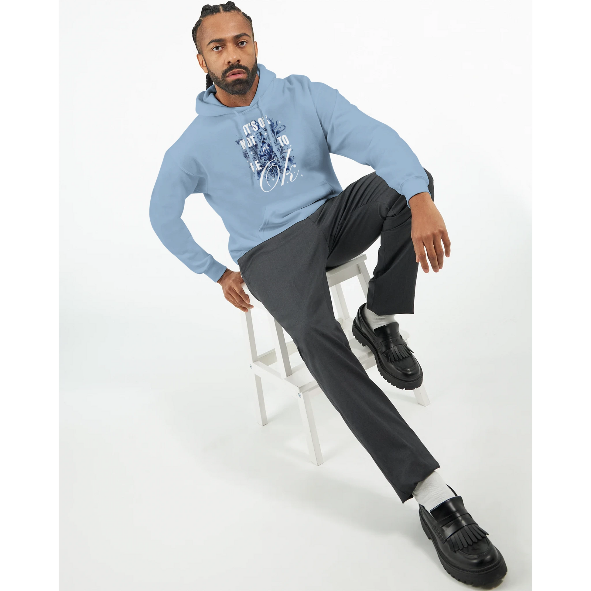 Man wearing light blue cotton-blend hoodie with 'It's OK Not To Be OK' graphic, seated on a white stool.