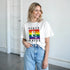 Woman wearing white Pride LGBTQ short-sleeve graphic tee with rainbow block and ampersand, Peace & Pride design.