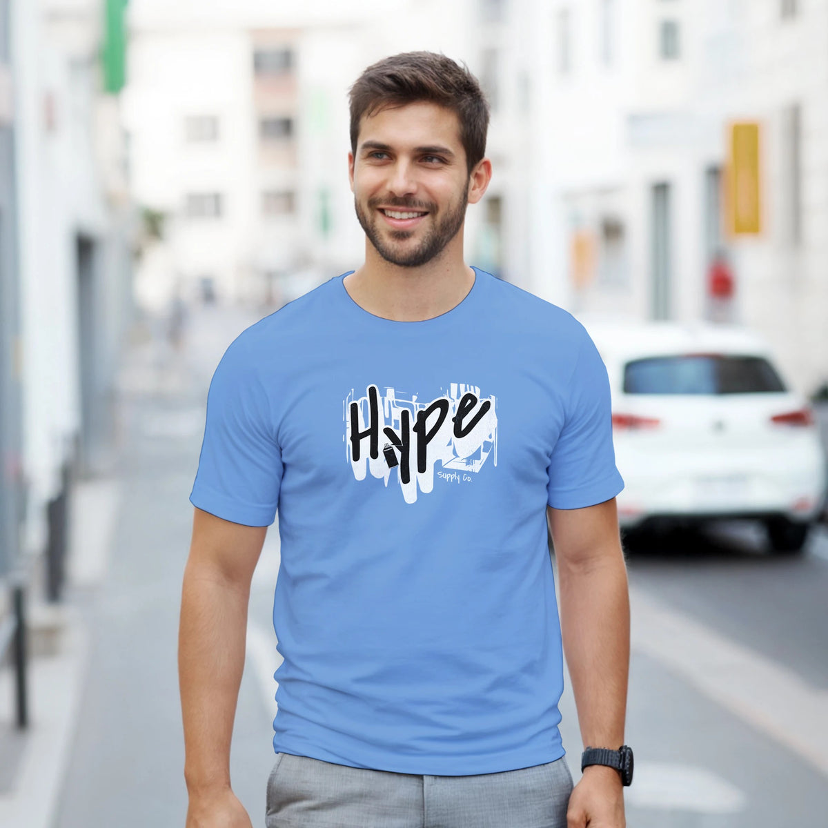 Man wearing Hype Men's Graphic T-Shirt, soft cotton blend blue tee with white brush-stroke logo on city street.