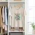 Beige t-shirt with floral design and 'Paradise' text hanging on a rack with other clothes.