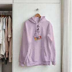Pastel pink retro cassette tape hoodie with unspooling tape graphic hanging on a wooden hanger in a closet.