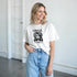 Woman wearing white Tropical Paradise graphic tee with blue and pink accents, soft cotton blend, light denim jeans.