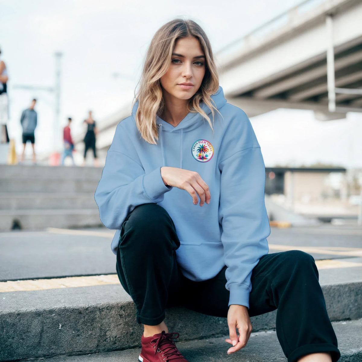 Woman crouching on steps in a light blue hoodie featuring a tropical palm tree graphic on the chest pocket.
