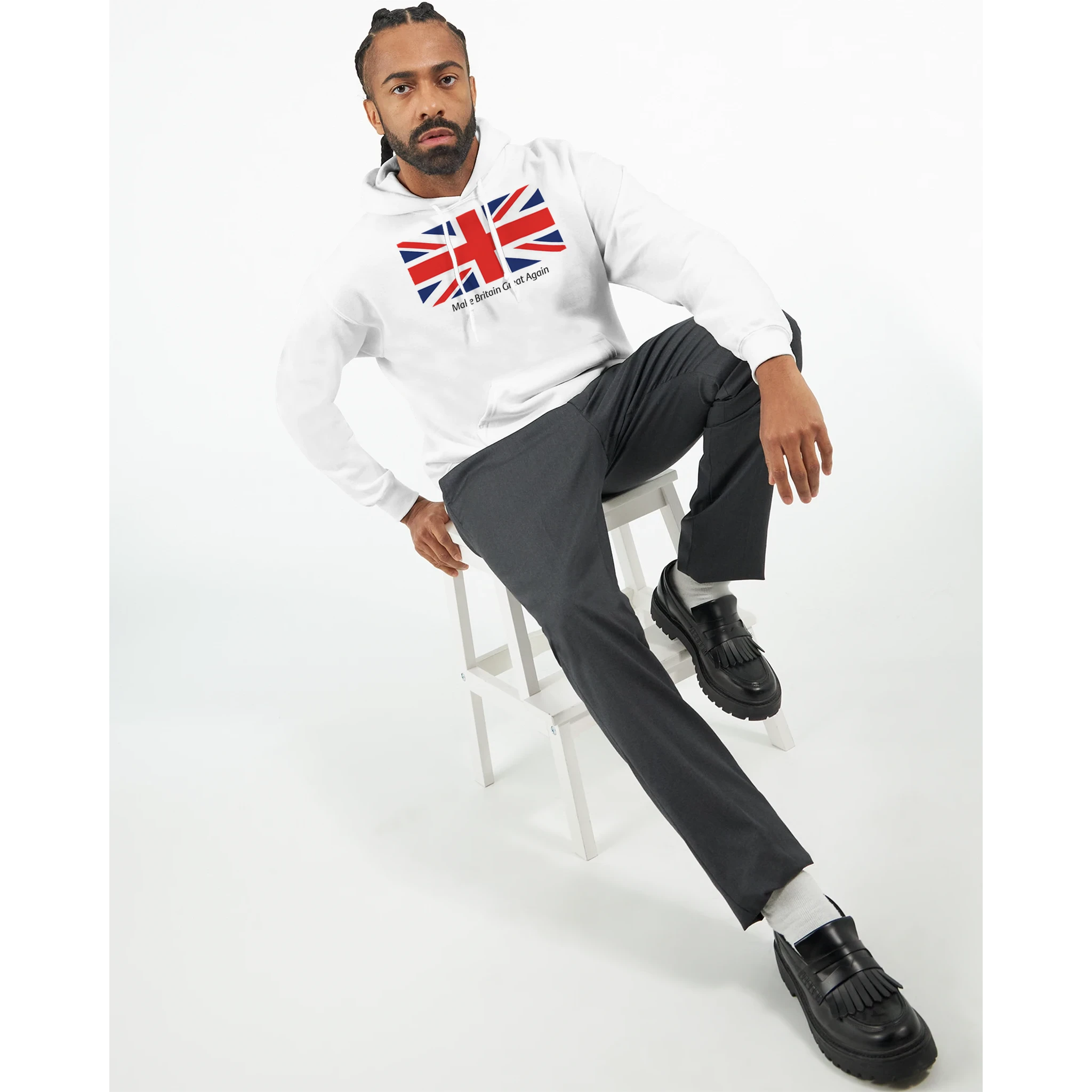 Man modeling a white Union Jack Hoodie with bold flag graphic, gray pants, black loafers, seated on a stool.