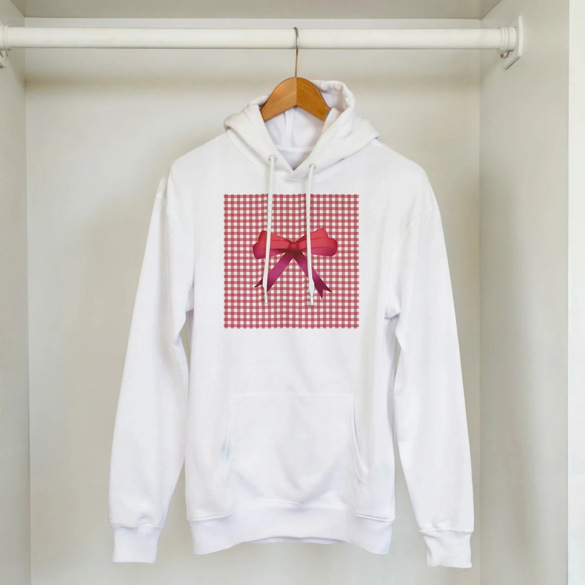 White hoodie with red gingham check blocks and a red bow graphic on front, hanging on a wooden hanger in a closet.