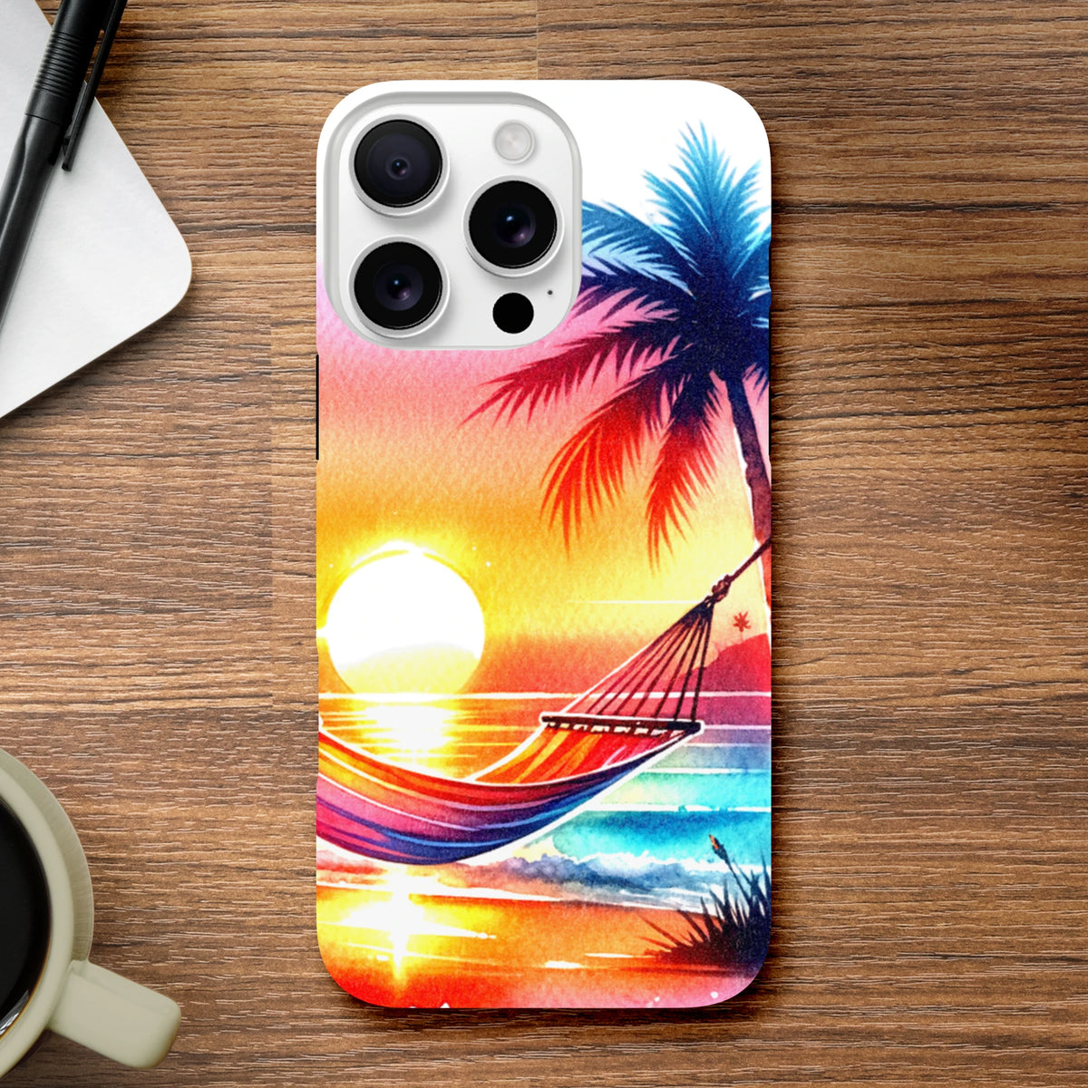 Vibrant tropical sunset phone case featuring a palm tree and hammock design on a white iPhone cover resting on a wooden desk.