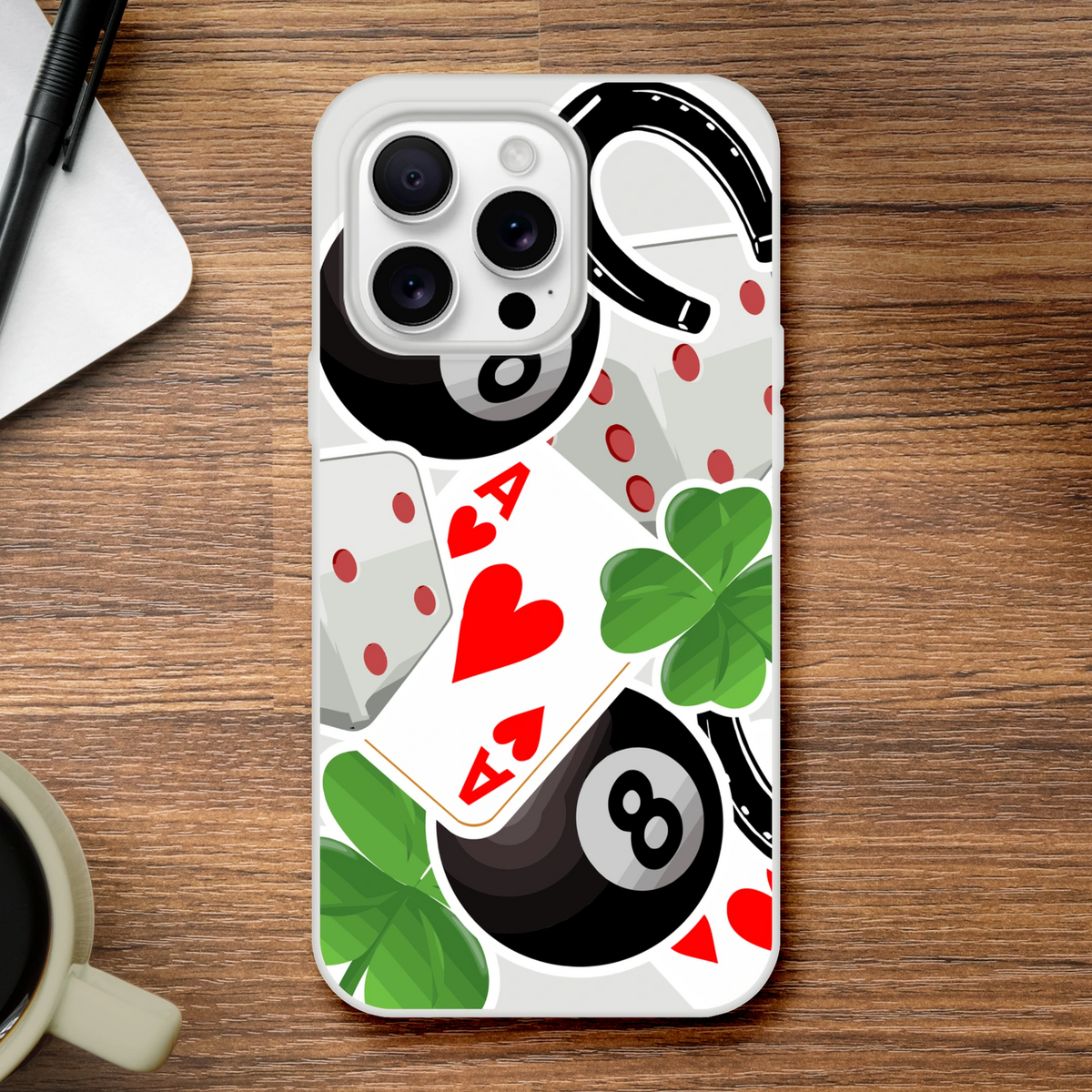 Clear iPhone case featuring lucky charm design with Ace of Hearts, 8 Ball, four-leaf clovers, and horseshoe on a wooden desk.