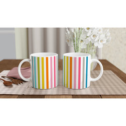 Pair of Rainbow Stripe Ceramic Mugs with bold vertical stripes on a wooden table beside white flowers.