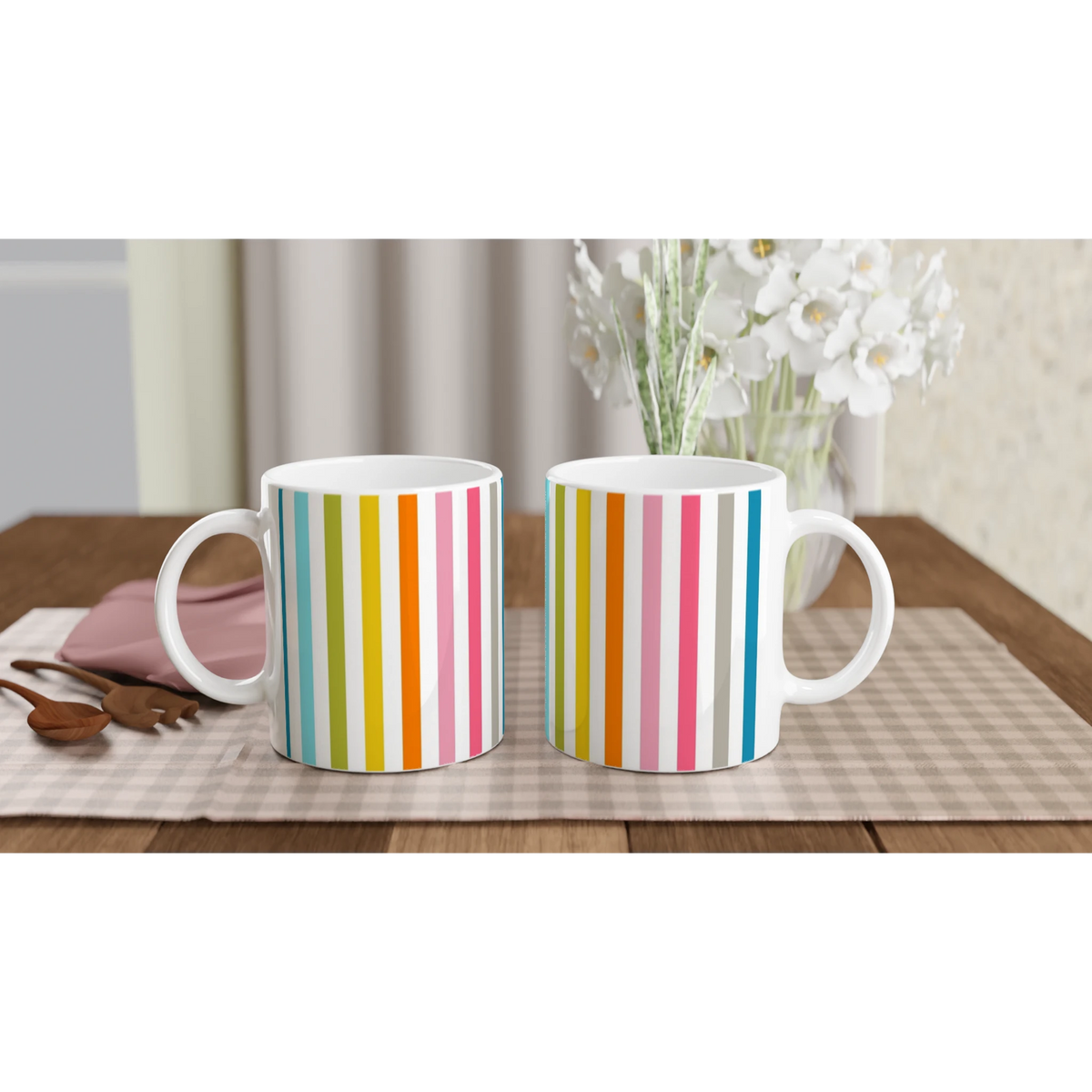 Pair of Rainbow Stripe Ceramic Mugs with bold vertical stripes on a wooden table beside white flowers.