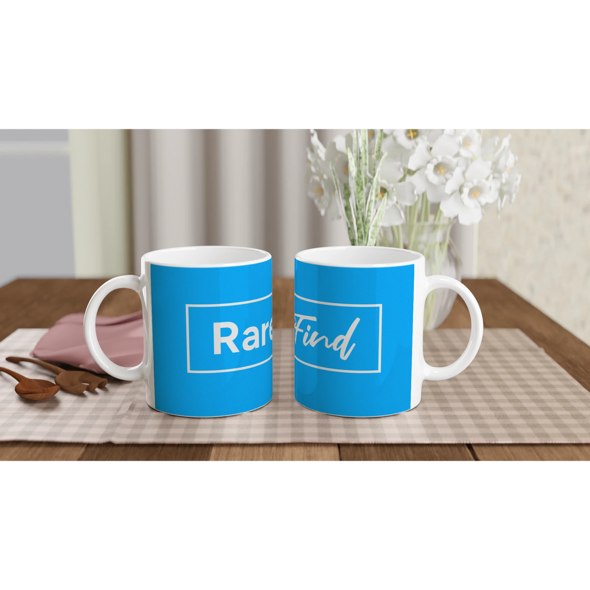 Two blue coffee mugs featuring Rare Find slogan on white handles, set on a table with white flowers in the background.