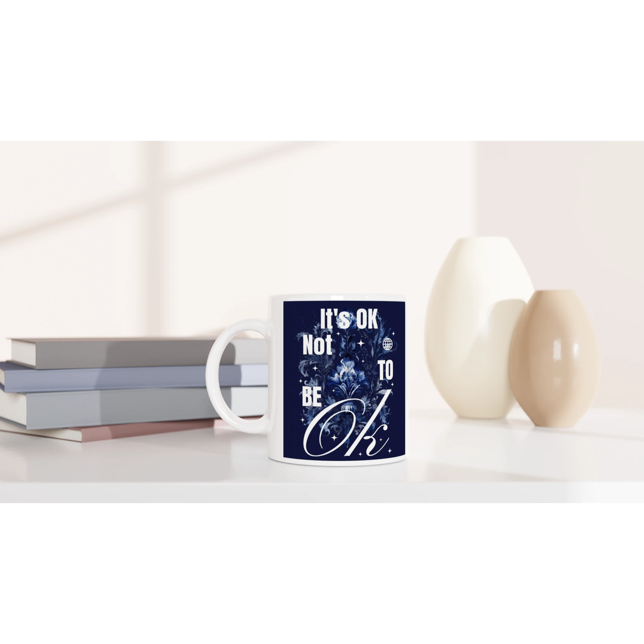 White ceramic mug with navy blue 'It's OK Not To Be OK' slogan and floral design, placed beside books.