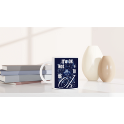 White ceramic mug with navy blue 'It's OK Not To Be OK' slogan and floral design, placed beside books.
