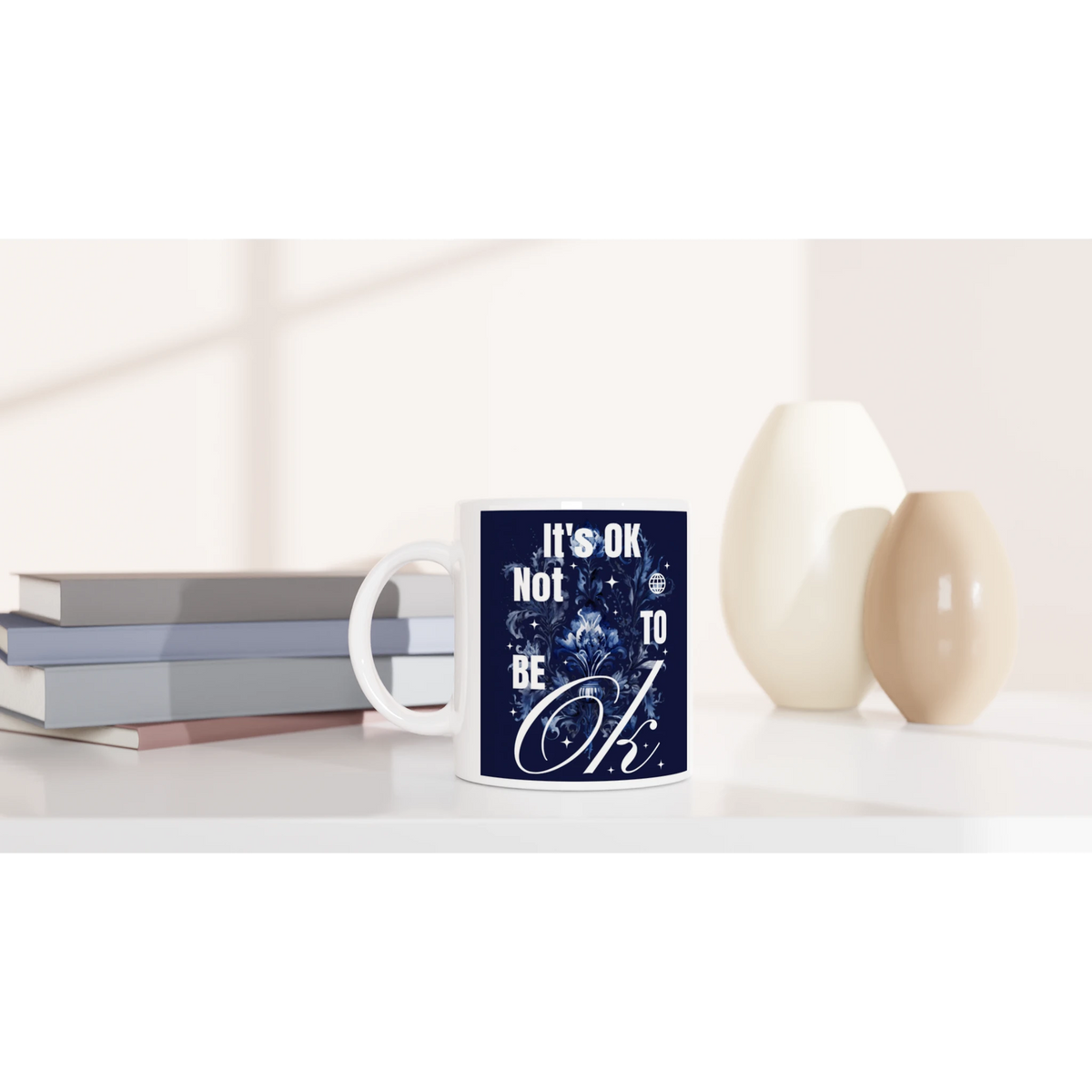 White ceramic mug with navy blue 'It's OK Not To Be OK' slogan and floral design, placed beside books.