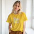 Woman in yellow Stay True T-shirt with white retro slogan, Urban Palm Trees streetwear vibe.