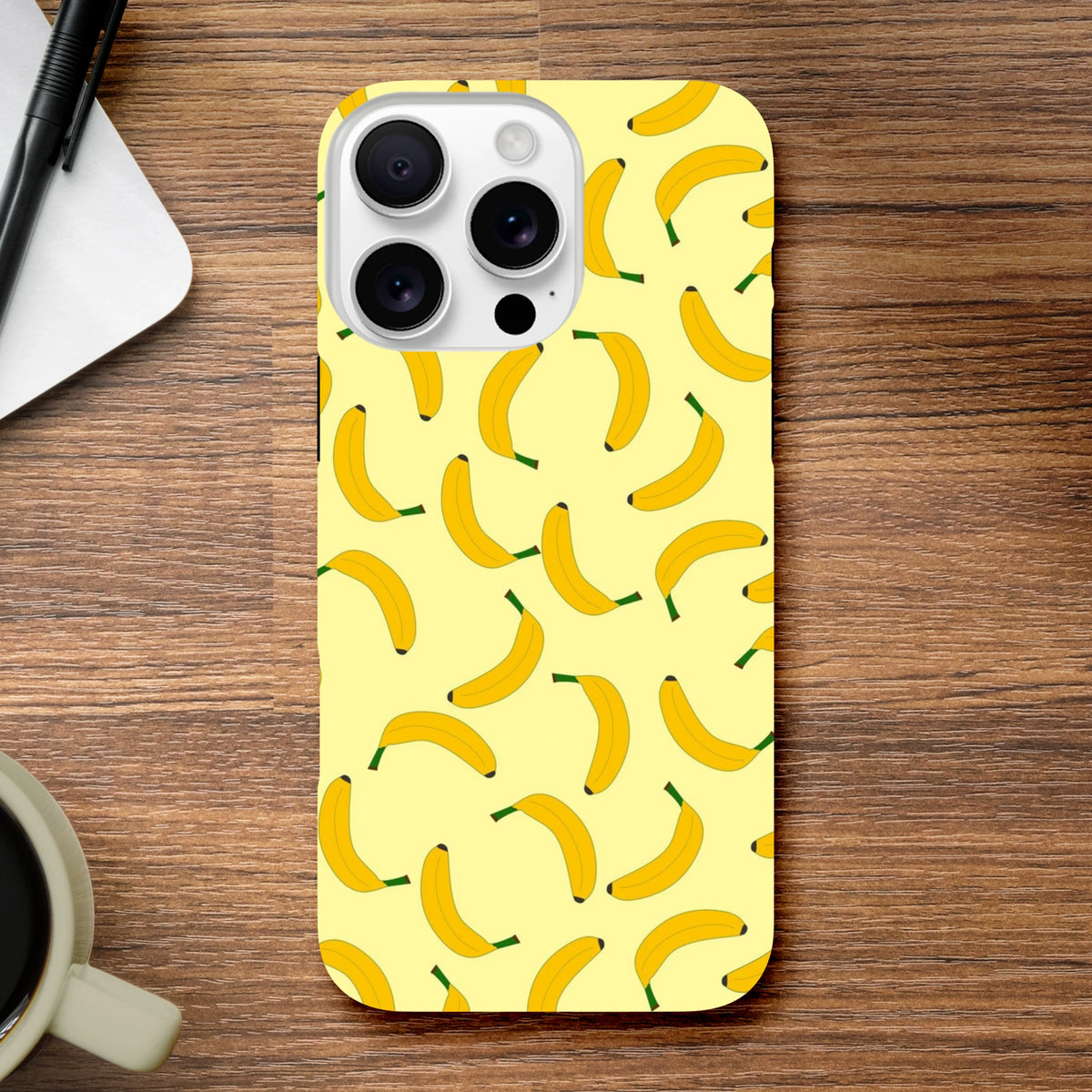 Yellow banana patterned tough iPhone case on a wooden desk. Cute fruit phone cover with playful energy and secure screen protection.
