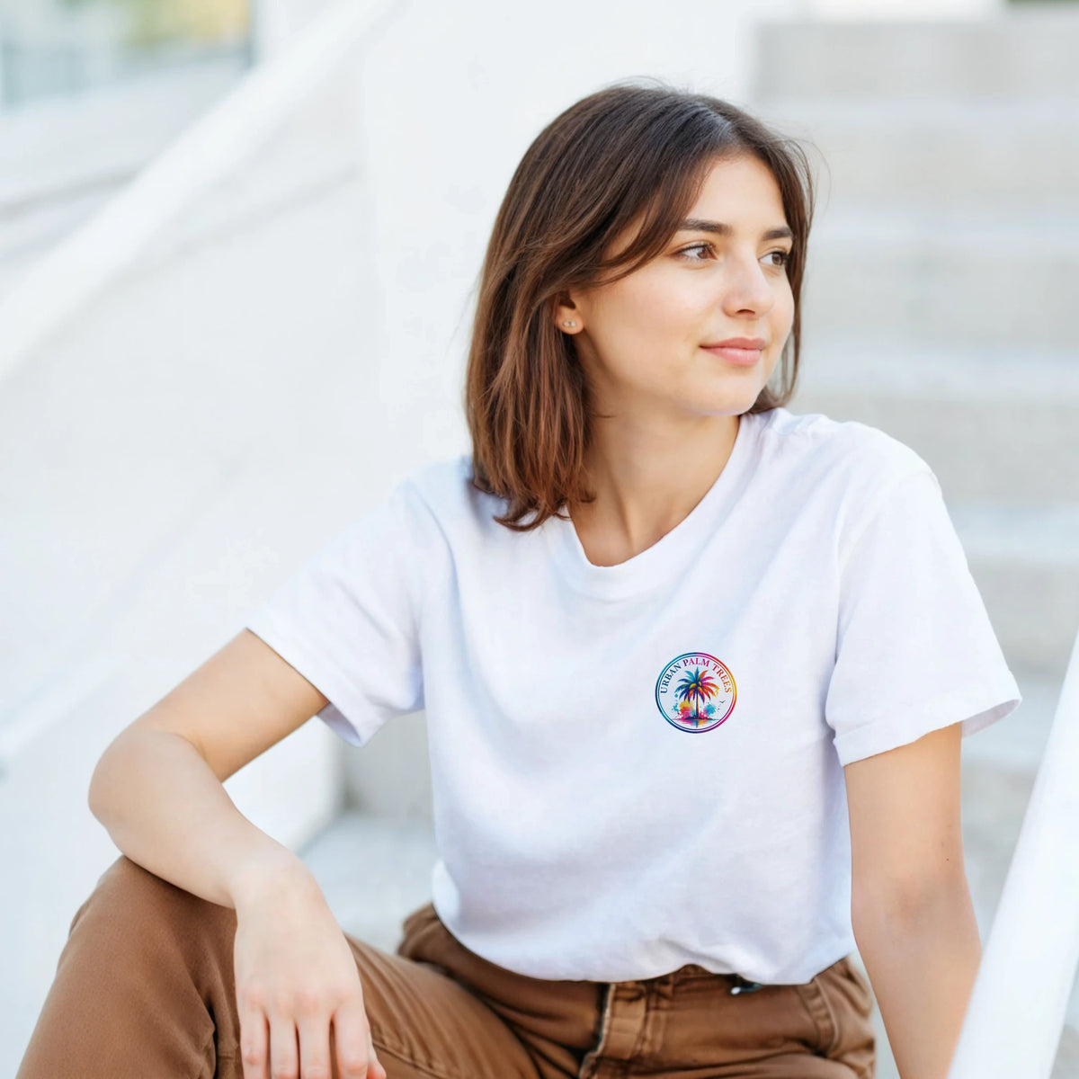 Woman wearing a white crew neck graphic tee with a small colorful circular palm tree logo on the chest.