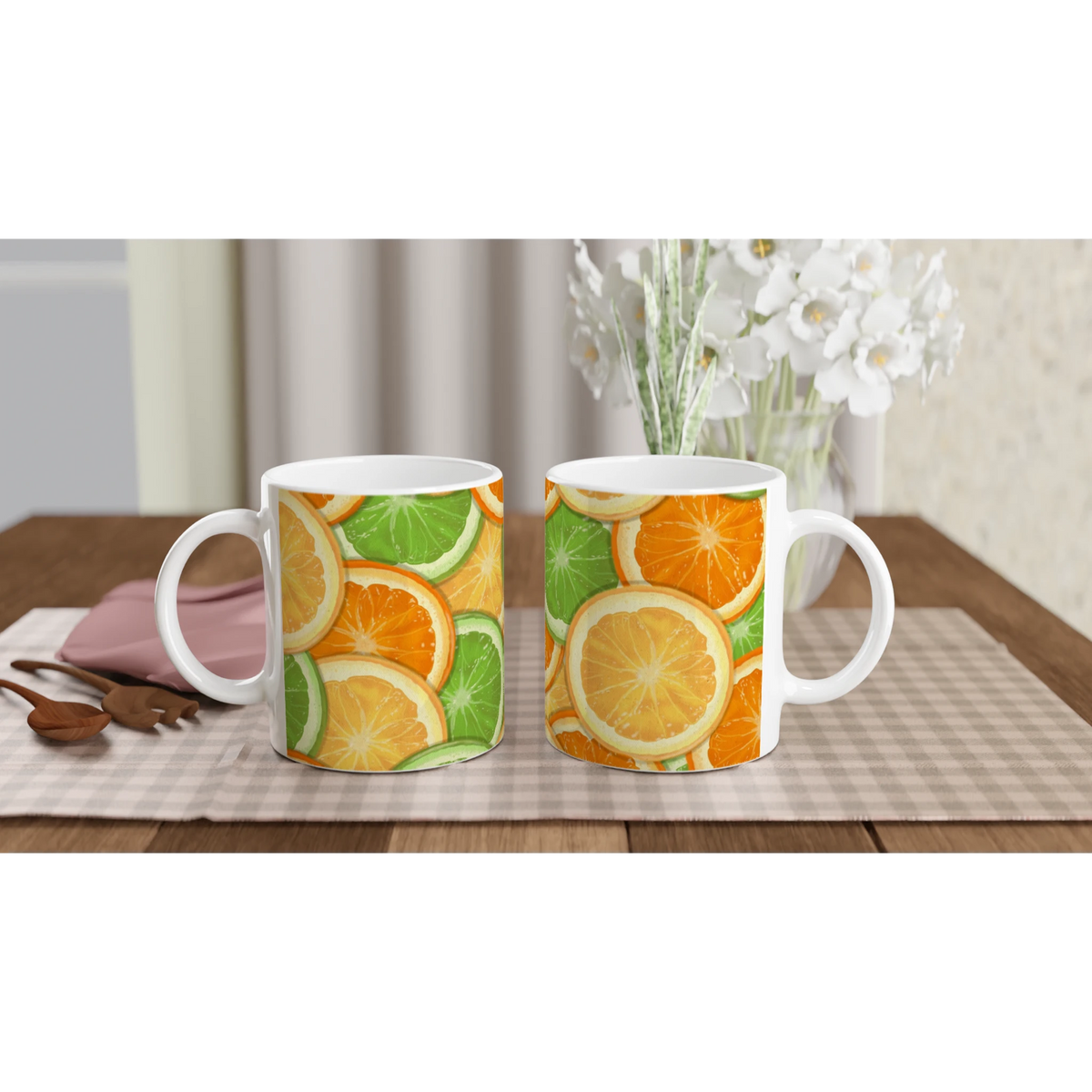Two citrus-pattern mugs with orange and lime slices on a checkered table near a vase of white flowers.