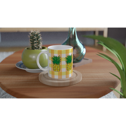 Pineapple-themed coffee mug with yellow gingham pattern on a wooden table beside a cactus.