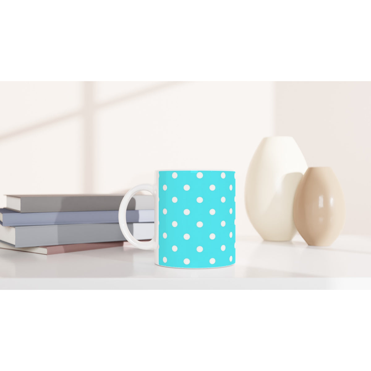 Aqua blue ceramic mug with white polka dots sits on a white desk next to stacked books and small vases.