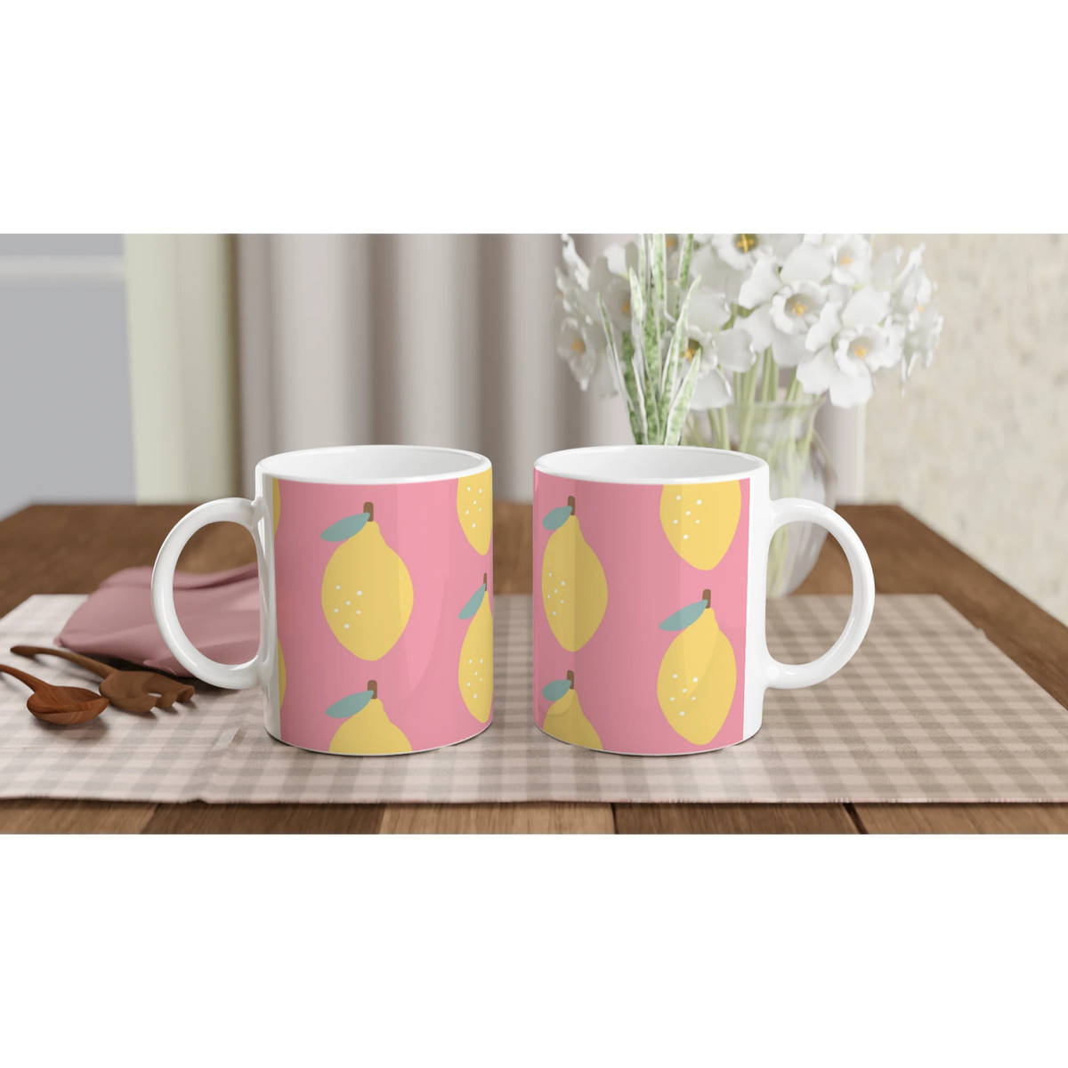 Two pink ceramic mugs with yellow lemon pattern on a checkered tablecloth and a floral vase in the background.