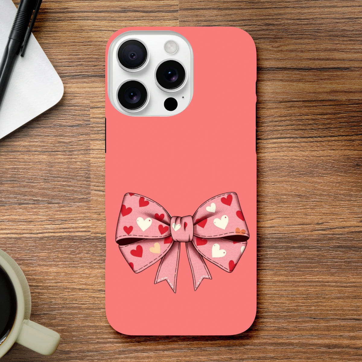 Pink iPhone case featuring a large bow pattern with red and white hearts on a coral background for cute coquette style.