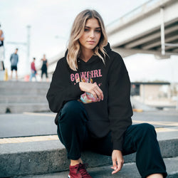 Woman in a black Cowgirl hoodie with pastel graphic, urban palm-tree backdrop.