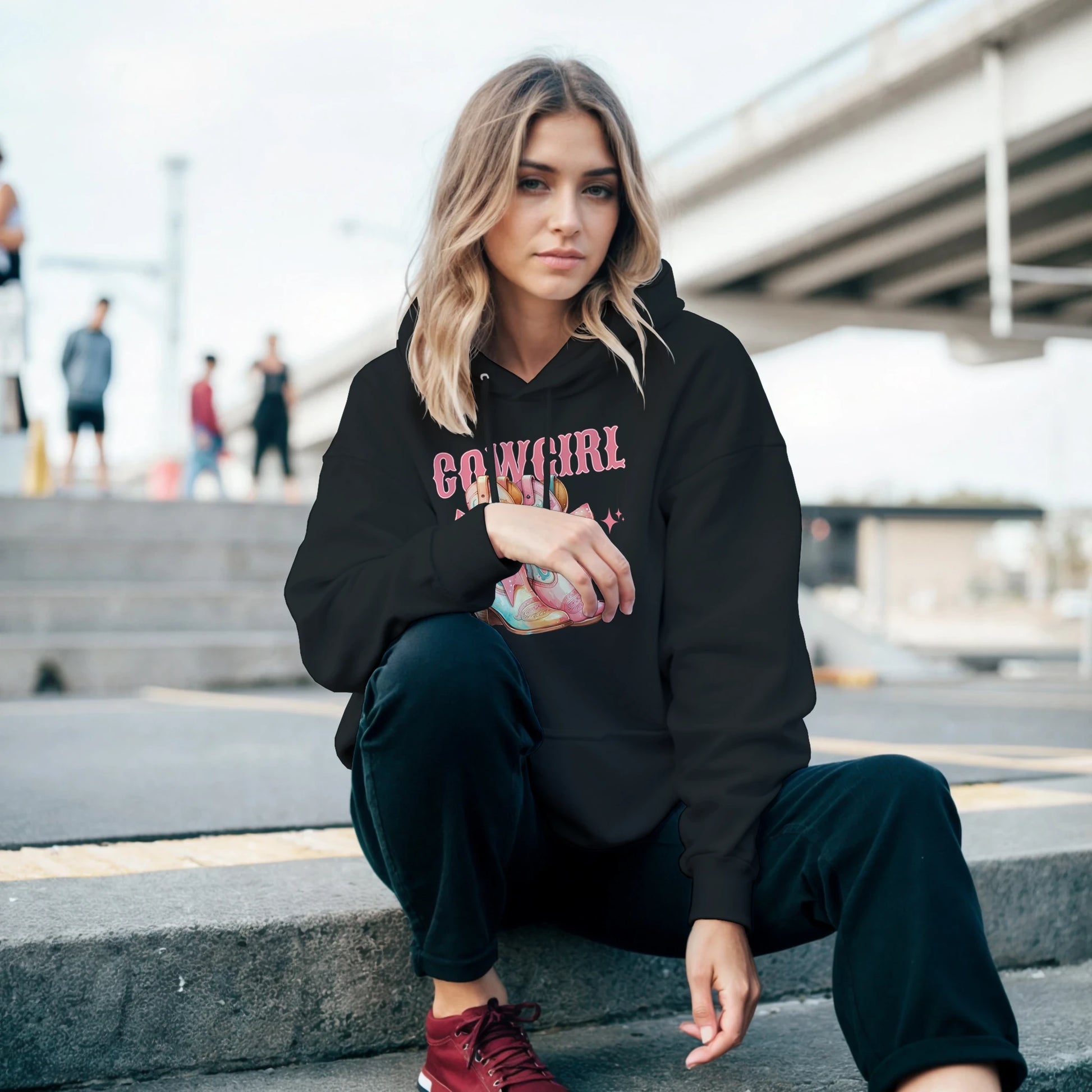 Woman in a black Cowgirl hoodie with pastel graphic, urban palm-tree backdrop.